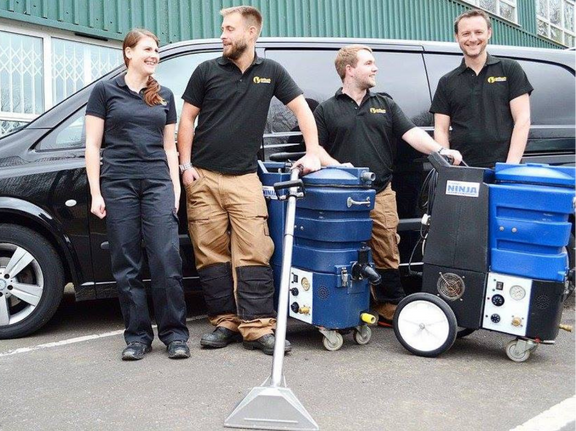 This is a photo of Buckhurst Hill Carpet Cleaning carpet cleaners (three men and one woman) standing in fromt of their black van, with two steam cleaning carpet machines next to them